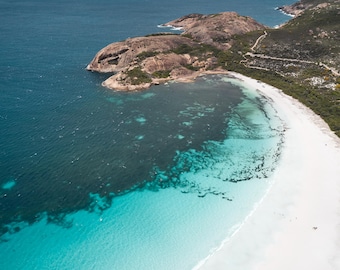 Könnte beinhalten: Eine Luftaufnahme eines weißen Sandstrandes, der auf türkisfarbenes und tiefblaues Meerwasser trifft. Eine felsige Landzunge mit braunen Gesteinsformationen ragt ins Meer, gesäumt von grüner Küstenvegetation und einem Parkplatz mit Fahrzeugen.