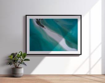 Könnte beinhalten: Gerahmter Kunstdruck mit einer Luftaufnahme einer türkisfarbenen und weißen Küstenlandschaft. Das Kunstwerk hängt an einer weißen Wand, mit einer kleinen Topfpflanze auf einer Holzoberfläche im Vordergrund. Der Rahmen ist schwarz.