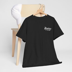 Peut inclure: T-shirt noir avec le texte "Anxiety is Everything" en blanc en police gothique. Une casquette de baseball blanche repose sur une chaise en bois en arrière-plan. Le t-shirt est suspendu à un cintre en bois.
