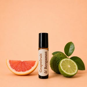 May include: A small amber glass roller bottle with a black cap and a white label reading Grapefruit & Bergamot. The bottle is positioned between a slice of pink grapefruit on the left and a whole and halved green bergamot fruit on the right.