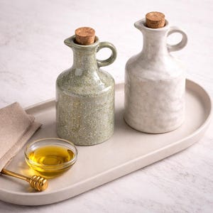 Puede incluir: Dos botellas de aceite de cerámica con tapones de corcho, una verde y otra blanca, sobre una bandeja crema. Un pequeño cuenco de cristal con miel y un cucharón de miel dorado también están en la bandeja, junto a un paño beige doblado. Las botellas tienen asas pequeñas.
