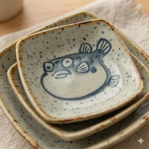 Puede incluir: Juego de tres platos cuadrados de cerámica con fondo moteado blanquecino y una ilustración de pez globo azul oscuro. Los bordes son de color marrón cálido. Adecuados para servir aperitivos o pequeñas porciones.