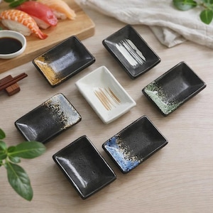 May include: A collection of rectangular ceramic soy sauce dishes. The dishes are primarily black with speckled accents of gold, white, blue, and green. One dish is white with gold brushstrokes. Sushi and chopsticks are in the background.