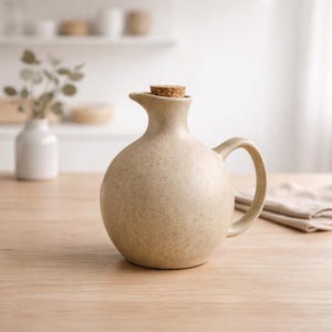 May include: A beige ceramic jug with a rounded body, a handle, and a spout. A cork stopper seals the top. The jug is on a wooden surface, with a white vase and greenery in the background. A neutral-coloured cloth is also visible.