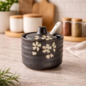 Puede incluir: Un azucarero de cerámica negra con tapa y una cuchara pequeña. El cuenco tiene una superficie texturizada y está decorado con diseños florales blancos. La cuchara es blanca. El fondo incluye otros artículos de cocina.
