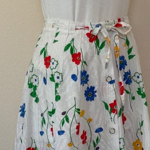May include: White wrap skirt with a floral pattern featuring red, blue, and yellow flowers. The skirt has a tie closure and button details. The flowers are set against a white background with green stems and leaves.