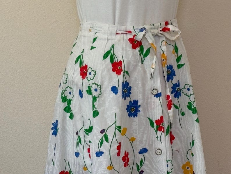May include: White wrap skirt with a floral pattern featuring red, blue, and yellow flowers. The skirt has a tie closure and button details. The flowers are set against a white background with green stems and leaves.