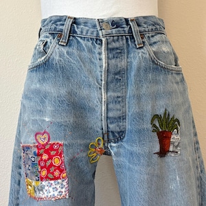 May include: Light blue denim jeans with decorative patches and embroidery. Patches include a red floral design and a patchwork design. Embroidery includes a yellow flower and a potted plant. The jeans have a button fly and classic five-pocket styling.