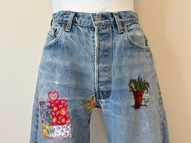 May include: Light blue denim jeans with decorative patches and embroidery. Patches include a red floral design and a patchwork design. Embroidery includes a yellow flower and a potted plant. The jeans have a button fly and classic five-pocket styling.