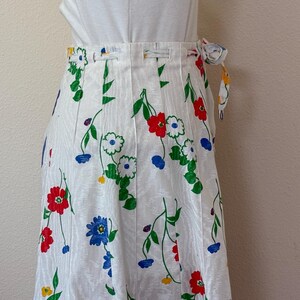 May include: White wrap skirt with a floral pattern featuring red, blue, and white flowers with green stems and leaves. The skirt has a tie closure and a vertical striped texture.