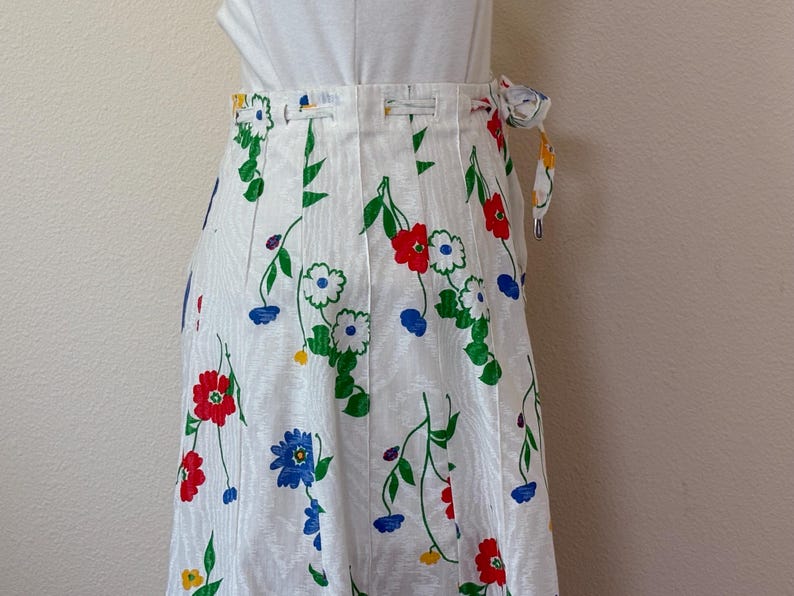 May include: White wrap skirt with a floral pattern featuring red, blue, and white flowers with green stems and leaves. The skirt has a tie closure and a vertical striped texture.
