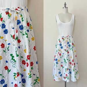 May include: White button-down skirt with a colorful floral pattern. The skirt features red, blue, and yellow flowers with green stems and leaves. It has a matching fabric belt and is paired with a white tank top.