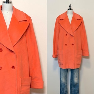 May include: A bright orange double-breasted coat featuring wide lapels, long sleeves with cuffs, and two large patch pockets on the front. The coat is fastened with matching orange buttons and is displayed on a mannequin wearing blue jeans.