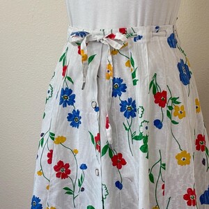 May include: White skirt with a colorful floral pattern featuring red, blue, and yellow flowers. The skirt has a tie-waist design and a button closure. The skirt is made of a lightweight fabric, perfect for a summer outfit.
