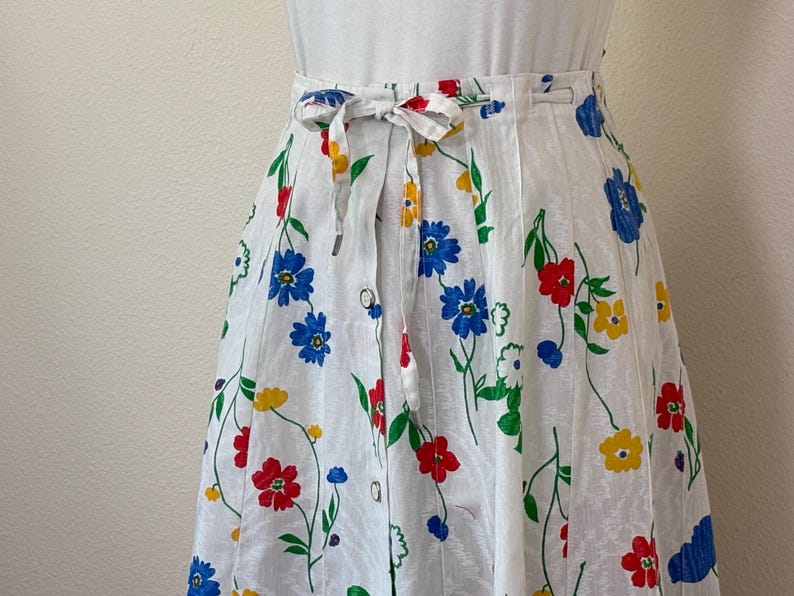May include: White skirt with a colorful floral pattern featuring red, blue, and yellow flowers. The skirt has a tie-waist design and a button closure. The skirt is made of a lightweight fabric, perfect for a summer outfit.