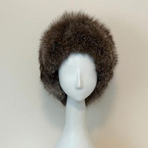 May include: A brown fur hat with a rounded crown. The hat is displayed on a white mannequin head against a plain background. The fur appears to be a mix of brown and grey.