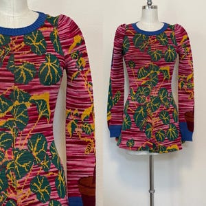 Può includere: Un top in maglia a maniche lunghe con un motivo botanico. Il top presenta uno sfondo a righe rosa e rosse con grandi disegni di foglie verdi e gialle. I bordini blu accentuano lo scollo e i polsini. L'indumento è esposto su un manichino.