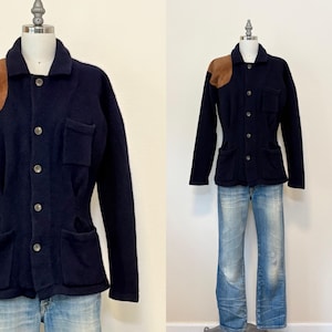 Puede incluir: Chaqueta azul marino con parches marrones en los hombros, cierre de botones y dos bolsillos delanteros. La chaqueta se exhibe en un maniquí con vaqueros azul claro. La chaqueta es de estilo vintage.