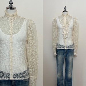 May include: Cream-colored lace blouse with a high ruffled collar and button-down front. The long sleeves have a slight puff at the shoulder and fitted cuffs. The blouse is paired with blue jeans.
