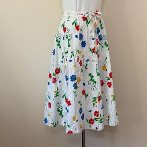 May include: A white wrap skirt with a colorful floral pattern. The skirt features red, blue, yellow, and green flowers with green leaves. It has a tie closure at the waist and is knee-length.