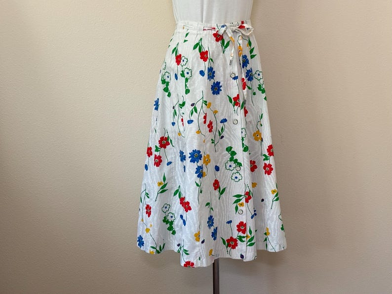 May include: A white wrap skirt with a colorful floral pattern. The skirt features red, blue, yellow, and green flowers with green leaves. It has a tie closure at the waist and is knee-length.