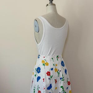 May include: A white tank top paired with a white skirt adorned with a colorful floral pattern. The skirt features red, yellow, and blue flowers with green stems and leaves. The outfit is displayed on a mannequin.