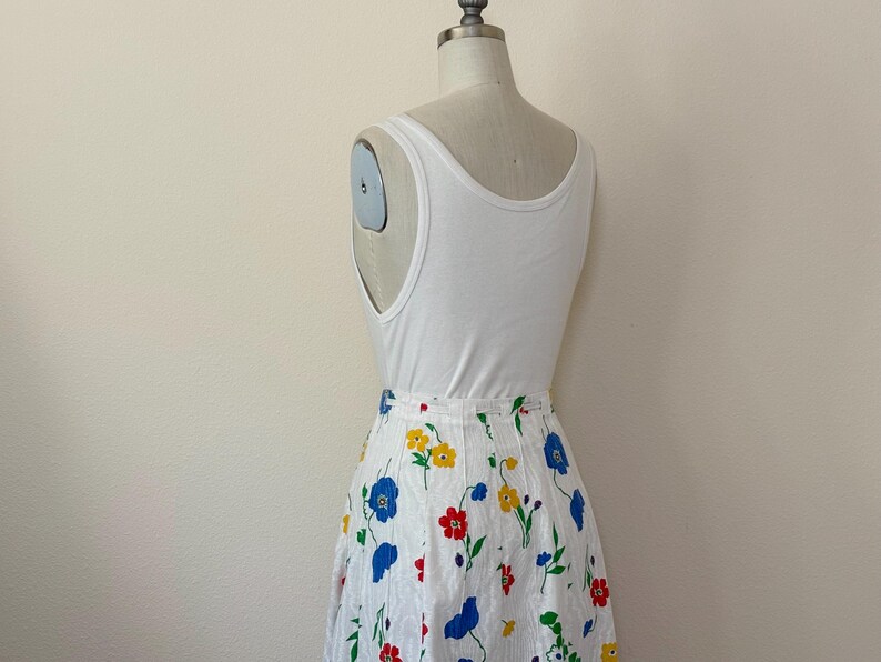 May include: A white tank top paired with a white skirt adorned with a colorful floral pattern. The skirt features red, yellow, and blue flowers with green stems and leaves. The outfit is displayed on a mannequin.