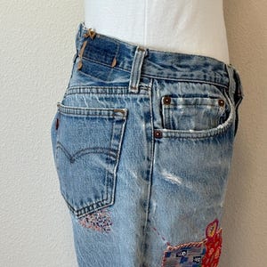 May include: Faded light blue denim jeans with decorative patches in red, blue, and pink. The jeans feature a classic five-pocket design, a button and zip fastening, and a distressed appearance. The Levi's logo is visible on the back pocket.