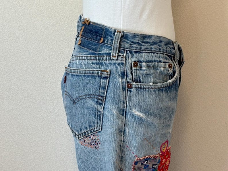 May include: Faded light blue denim jeans with decorative patches in red, blue, and pink. The jeans feature a classic five-pocket design, a button and zip fastening, and a distressed appearance. The Levi's logo is visible on the back pocket.