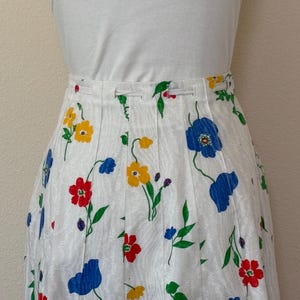 May include: White pleated skirt with a colorful floral pattern. The skirt features red, yellow, and blue flowers with green stems and leaves. The skirt is paired with a white top.