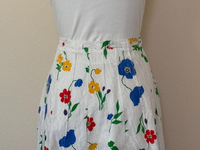 May include: White pleated skirt with a colorful floral pattern. The skirt features red, yellow, and blue flowers with green stems and leaves. The skirt is paired with a white top.