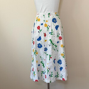 May include: A white skirt with a floral pattern featuring red, blue, and yellow flowers. The skirt has a high waist and a knee-length hemline. The fabric has a subtle textured appearance.
