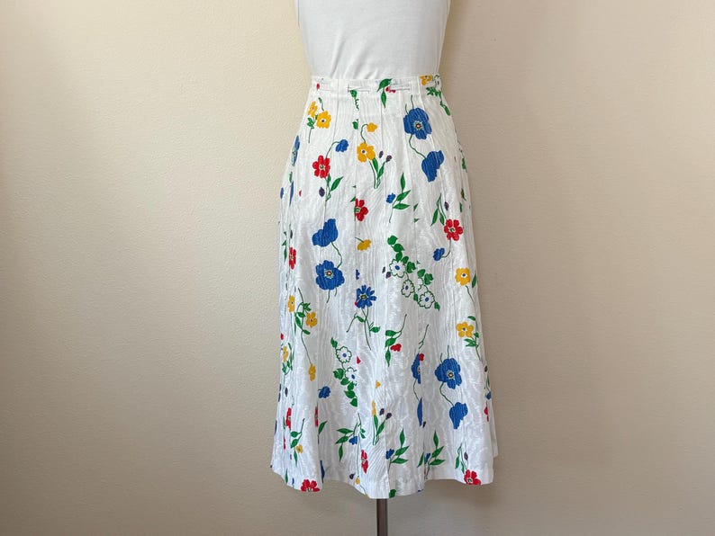 May include: A white skirt with a floral pattern featuring red, blue, and yellow flowers. The skirt has a high waist and a knee-length hemline. The fabric has a subtle textured appearance.