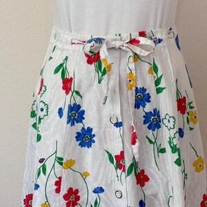 May include: White floral skirt with a button-down front and a tie waist. The skirt features a colorful floral pattern with red, blue, and yellow flowers and green stems. The skirt is made of a lightweight fabric.