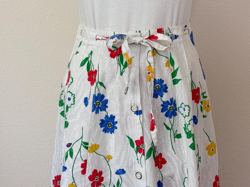 May include: White floral skirt with a button-down front and a tie waist. The skirt features a colorful floral pattern with red, blue, and yellow flowers and green stems. The skirt is made of a lightweight fabric.