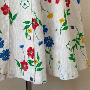 May include: White fabric with a floral pattern featuring red, blue, and yellow flowers with green leaves. The fabric is buttoned down the front. The flowers are in a repeating pattern.