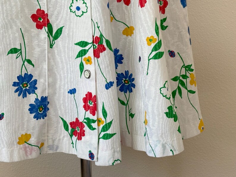 May include: White fabric with a floral pattern featuring red, blue, and yellow flowers with green leaves. The fabric is buttoned down the front. The flowers are in a repeating pattern.