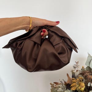 May include: A dark brown satin handbag featuring a gathered, ruched design with a soft, rounded silhouette and a knotted top handle. The fabric has a subtle sheen, creating deep folds and shadows across the surface of the pouch-style bag.