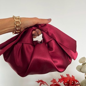 Puede incluir: Un bolso de mano de satén color burdeos intenso con un diseño de asa anudada y una textura de tela suave y fruncida. El bolso tiene un acabado liso y brillante y una silueta redondeada y holgada que crea pliegues en la superficie.
