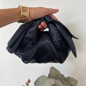 Könnte beinhalten: Eine dunkelmarineblaue Knotentasche aus Satin mit weicher, geraffter Stoffkonstruktion und einem geknoteten Griffdesign. Die Tasche hat eine glatte, glänzende Oberfläche und eine lockere, abgerundete Form. Ein breiter, geometrischer Armreif aus goldfarbenem Metall ist sichtbar.