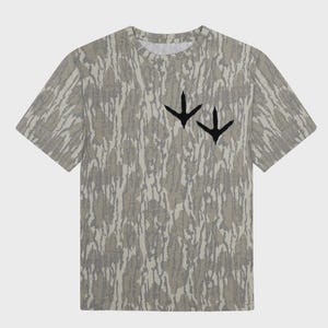 Puede incluir: Una camiseta de manga corta con un estampado de camuflaje en tonos grises y beige. La camiseta presenta dos huellas de p&aacute;jaros negras en el pecho. Tiene cuello redondo y un corte holgado.