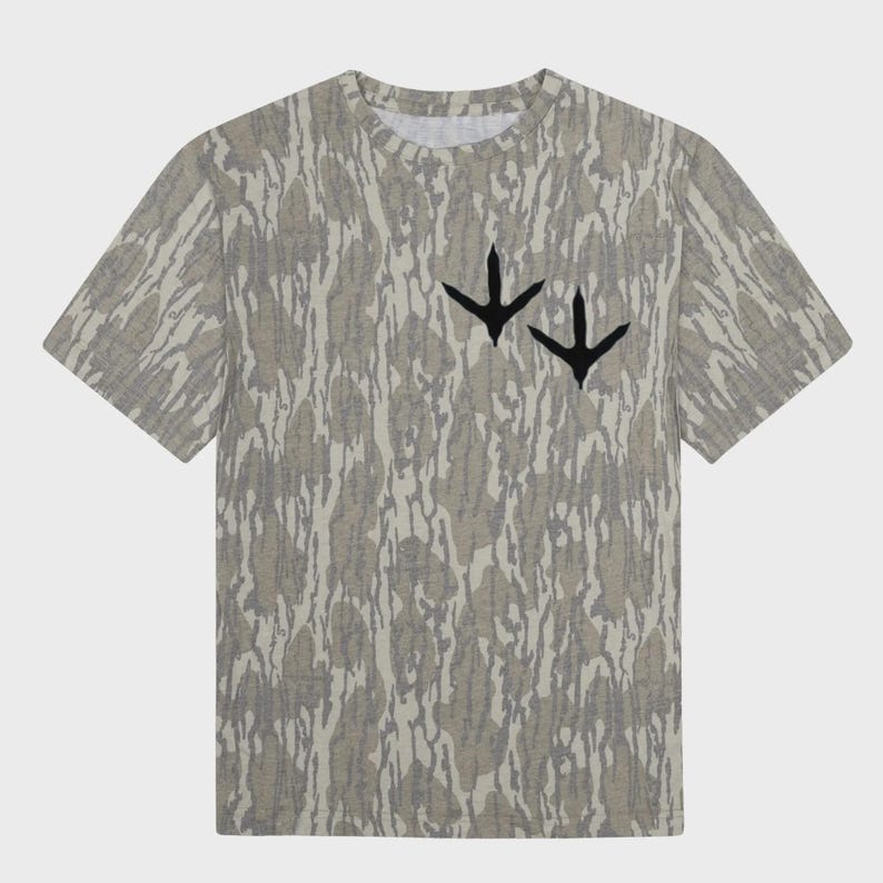 Puede incluir: Una camiseta de manga corta con un estampado de camuflaje en tonos grises y beige. La camiseta presenta dos huellas de p&aacute;jaros negras en el pecho. Tiene cuello redondo y un corte holgado.