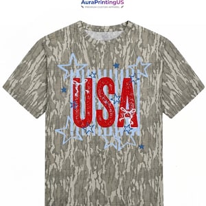 May include: A short-sleeve t-shirt with a camouflage pattern in shades of brown, tan, and green. The center features the red text USA with a distressed texture, surrounded by light blue outlined stars and small white ribbon bow accents.