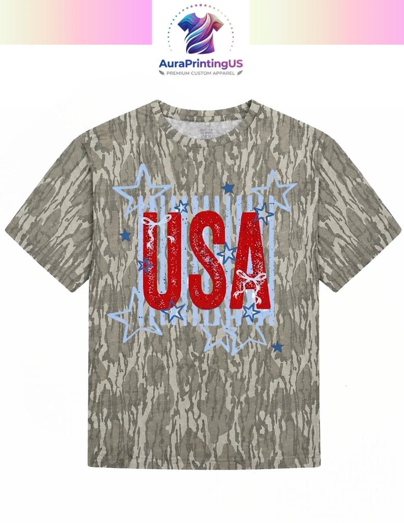 May include: A short-sleeve t-shirt with a camouflage pattern in shades of brown, tan, and green. The center features the red text USA with a distressed texture, surrounded by light blue outlined stars and small white ribbon bow accents.