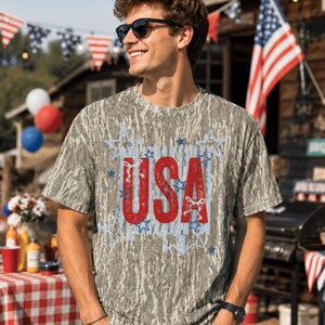 May include: A short-sleeve t-shirt with a gray and brown tree bark camouflage pattern. The center of the shirt features the text USA in large, distressed red letters, surrounded by a light blue graphic outline of multiple stars.