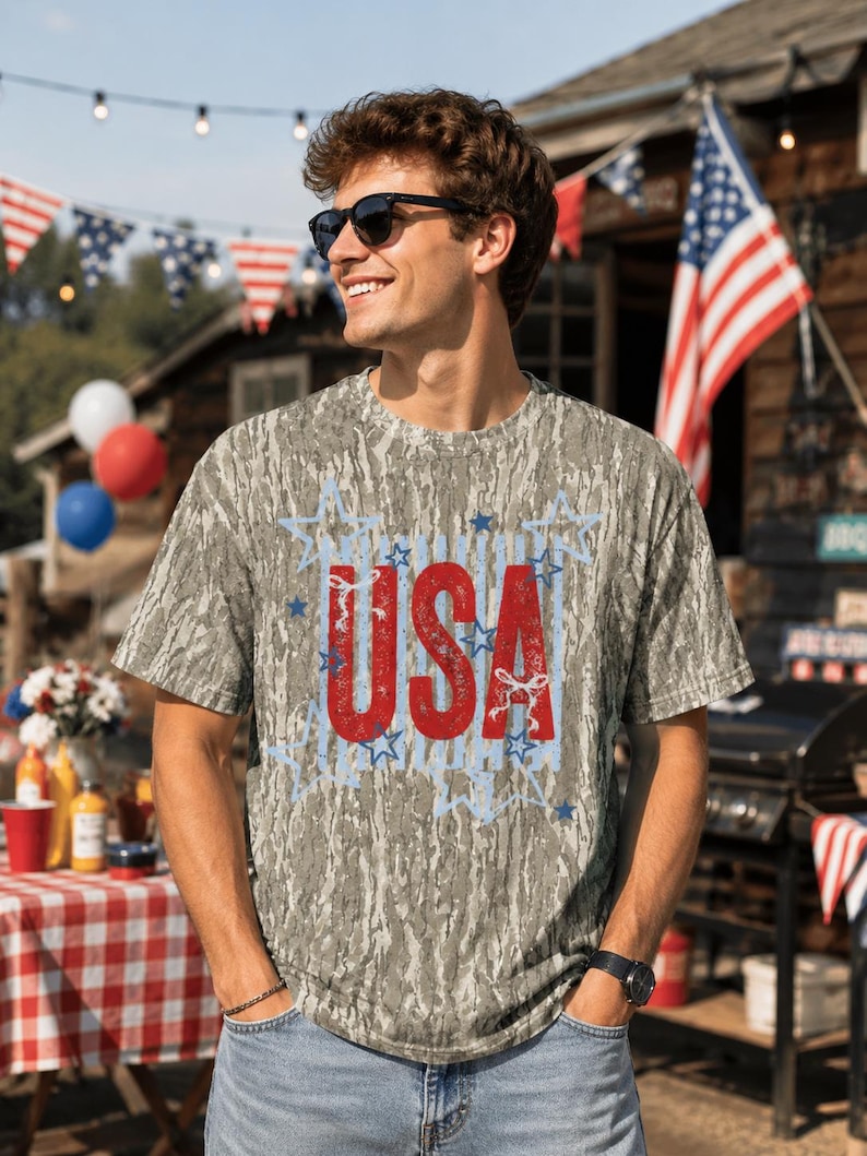 May include: A short-sleeve t-shirt with a gray and brown tree bark camouflage pattern. The center of the shirt features the text USA in large, distressed red letters, surrounded by a light blue graphic outline of multiple stars.