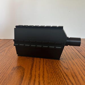 Pode incluir: Um objeto retangular preto com uma extensão cilíndrica no lado direito. A parte superior e as laterais têm um design com nervuras. O objeto é feito de um material preto fosco e está sobre uma superfície de madeira.
