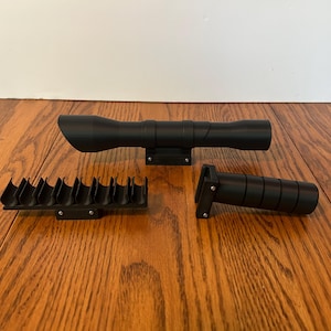 Op de afbeelding: Drie zwarte vuurwapenaccessoires worden weergegeven op een houten oppervlak. De items omvatten een richtkijker, een handgreep en een magazijnhouder. De richtkijker is cilindrisch met een uitlopend uiteinde. De handgreep is ook cilindrisch.