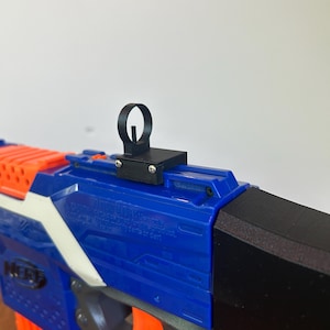 May include: A black sight attachment is mounted on a blue and orange toy gun. The toy gun has a blue body with white accents and orange details. The sight is a small, black, loop-shaped component.