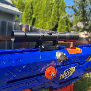 May include: A black cylindrical toy scope mounted on top of a blue and orange plastic toy blaster. The blaster features the yellow NERF logo on the side, with an orange circular attachment point and a small metallic silver circular detail near the scope.